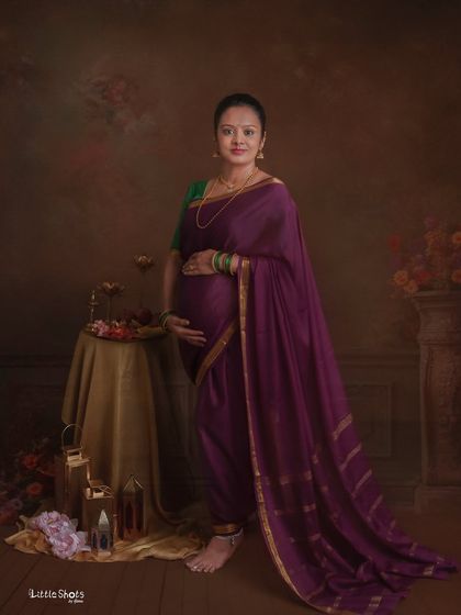 A full-length portrait of a mother-to-be in a traditional purple saree. She stands gracefully in a studio with a classic, painterly backdrop.
