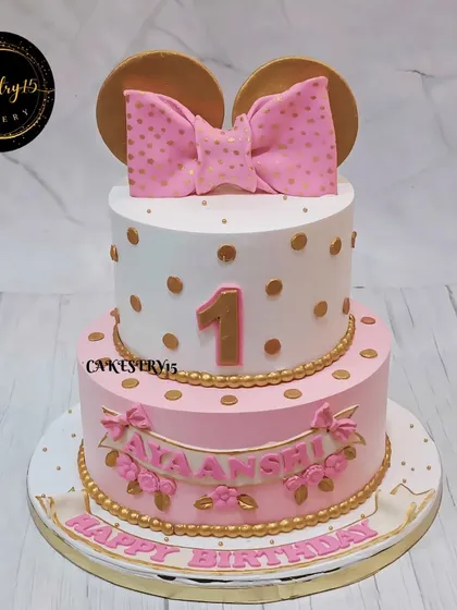 An enchanting two-tier Disney-themed cake for a first birthday. This chocolate cake features a large pink bow and golden polka dots, reminiscent of Minnie Mouse, for a classic and elegant look.