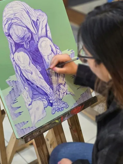 An over-the-shoulder view shows a student meticulously working on the lower portion of her figurative ballpoint pen artwork.