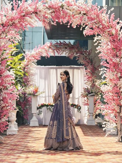A back view of the client in the mauve-purple lehenga, framed by the pink floral arch.