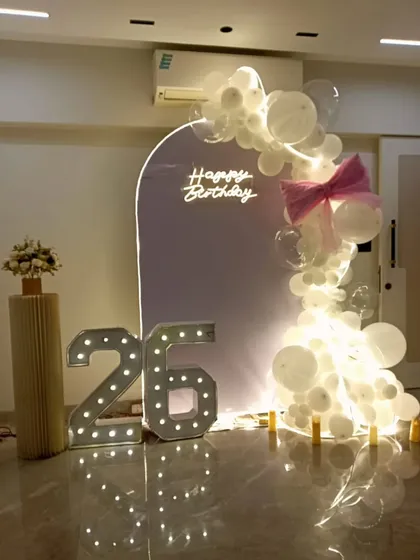 A minimalist and elegant setup for a 26th birthday. A simple arched backdrop with a neon sign is paired with a delicate white balloon garland and large marquee numbers.