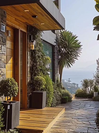 A modern entrance combining natural materials. We used a wooden deck for the porch, stacked stone for the wall, and a stone pathway, all softened by lush greenery in contemporary planters.