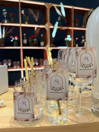 Elegant custom tags for a wedding, featuring the couple's initials and their hashtag, '#JasWeMetEachOther', clipped to the glassware.