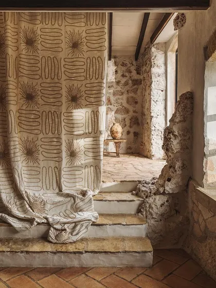 The "Perpétuel Soleil" fabric from Élitis, used as a curtain in a rustic stone interior. The large-scale, hand-beaded embroidery catches the light, transforming the drapery into a piece of textile art that bridges the gap between rustic architecture and refined design.