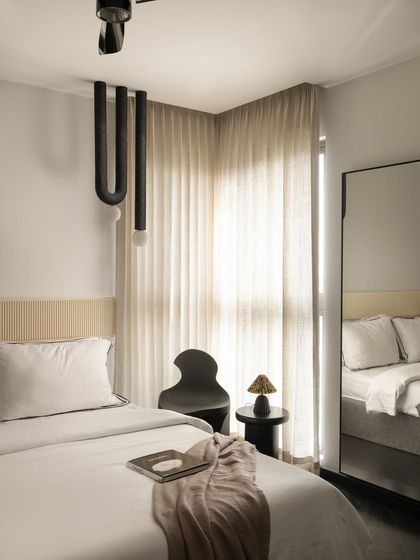 This guest bedroom at Ecru Maison features a minimalist design with a custom fluted headboard panel. A sculptural black chair and a unique U-shaped pendant light add artistic, modern touches to the calm space.