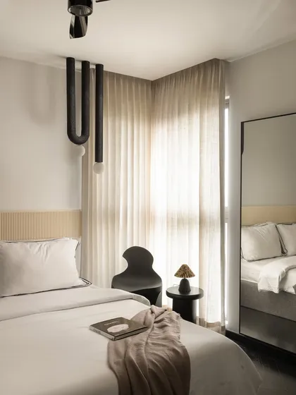 This guest bedroom at Ecru Maison features a minimalist design with a custom fluted headboard panel. A sculptural black chair and a unique U-shaped pendant light add artistic, modern touches to the calm space.