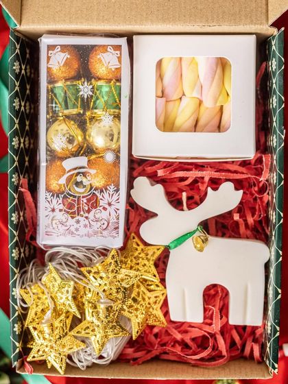 A whimsical Christmas gift box filled with holiday magic. It includes a reindeer-shaped candle, a string of star-shaped fairy lights, marshmallows, and festive chocolates.