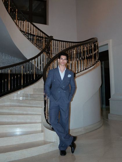 A full view of the grey double-breasted suit, showcasing its elegant drape and the client's confident posture. The luxurious setting complements the sophisticated tailoring.