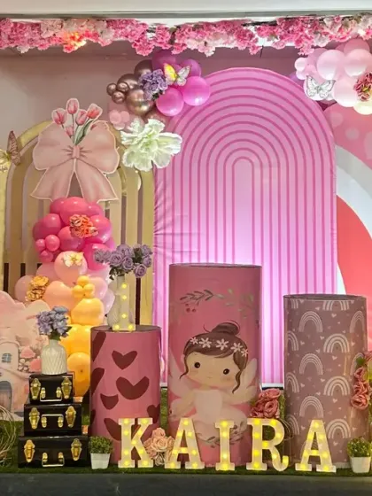 An enchanting fairy-themed first birthday party. This elaborate setup includes a large teddy bear, mushroom house props, custom-printed plinths, and a beautiful pink and gold balloon garland under a floral arch.