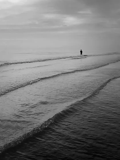 A solitary figure stands in the shallow waves, creating a sense of quiet contemplation. The repeating lines of the waves lead the eye through this calm and minimalist seascape.