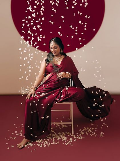 Seated gracefully amidst falling petals, she holds a peacock feather, a symbol of divinity and beauty. This artistic studio shot is a celebration of creation.