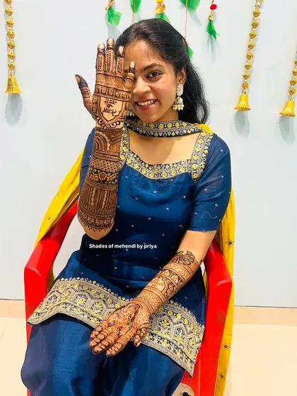 A happy bride showing off her intricate mehendi. One arm features a full bridal story, while the other has a portrait of a spiritual guru, combining personal and divine elements.