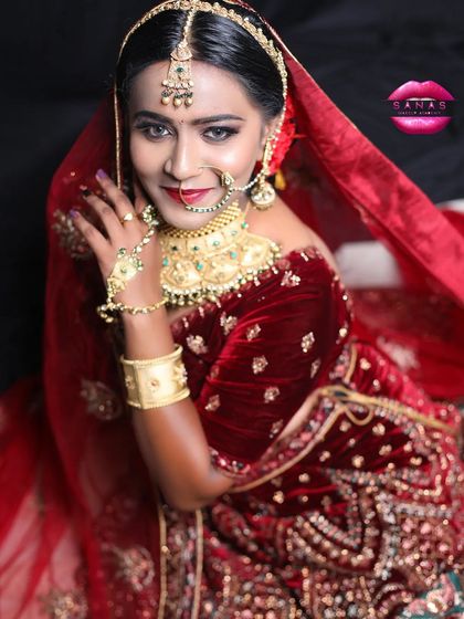 A royal bridal look featuring a deep red velvet lehenga. The makeup is classic and bold, with a red lip and defined eyes, complemented by a beautiful gold and green choker.