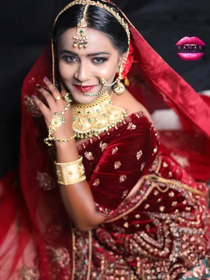 A royal bridal look featuring a deep red velvet lehenga. The makeup is classic and bold, with a red lip and defined eyes, complemented by a beautiful gold and green choker.