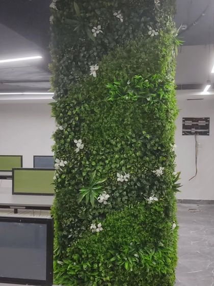 An office pillar wrapped in a dense artificial green panel with white flowers. This is a great way to hide structural elements and bring nature into an open-plan office.