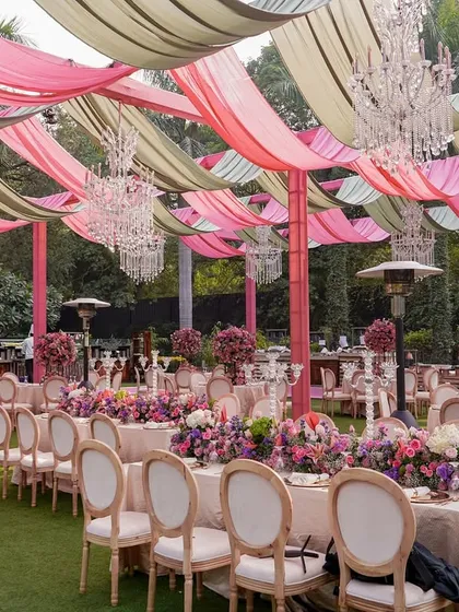 An elegant outdoor dining setup for a garden party. The tables are adorned with lush pink and purple floral runners, under a ceiling of draped fabric and crystal chandeliers.