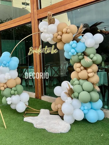 This outdoor bachelorette setup uses a gold ring frame as the base for a beautiful balloon arrangement. The earthy tones of sage green, beige, and light blue, along with dried pampas grass accents, create a lovely boho-chic look perfect for a garden party.
