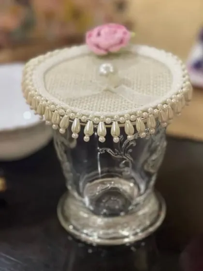 A close-up of a single embellished glass cover on an ornate glass. This piece features a delicate pink rose and a pearl-draped edge, perfect for protecting a drink at a garden party or bridal shower.
