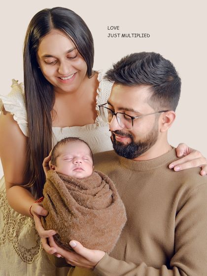 A warm, earth-toned family portrait. The parents look down at their swaddled newborn with pure affection, a simple and beautiful representation of their love multiplying.