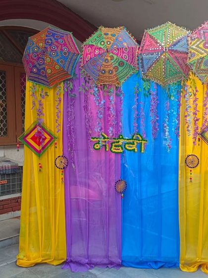 A vibrant Mehandi backdrop with drapes in yellow, purple, and blue. The decor is enhanced with hanging floral strings and colorful Rajasthani umbrellas for a festive look.