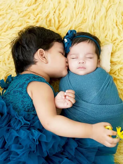 The sweet bond between sisters is a story in itself. Here, a big sister gently kisses her newborn sibling, both dressed in beautiful matching gowns. It's a quiet, tender moment that melts my heart.