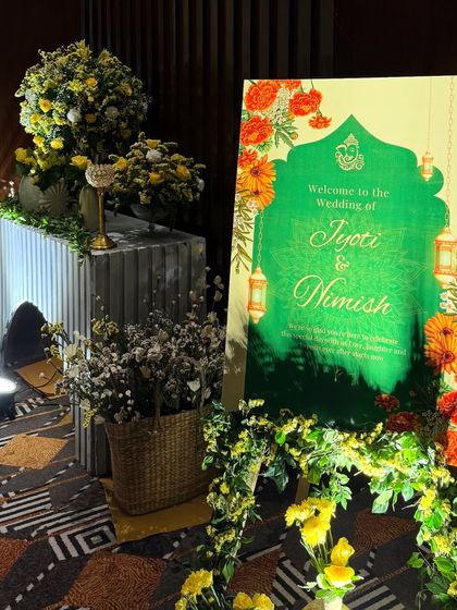 A personalized welcome to Jyoti and Nimish's wedding. The custom green welcome sign is framed by a garland of yellow flowers and surrounded by matching floral arrangements and rustic baskets.