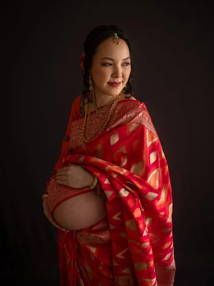 A striking maternity portrait focusing on the beauty of the bare bump against the rich fabric of a Banarasi saree. The dark background makes the colours and textures pop.