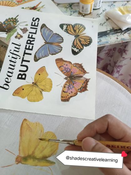 A student practices painting a butterfly on fabric, using a reference book for detail. Our classes emphasize learning foundational techniques, from color blending to fine brushwork.