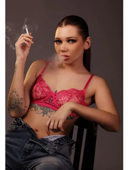 A defiant and edgy portrait of a model in a pink bra and jeans, smoking a cigarette against a grey studio background.