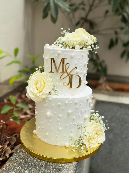 For this two-tier engagement cake, I went for a classic and elegant look. The design features delicate buttercream pearls, fresh white roses, baby's breath, and a custom gold acrylic monogram for the couple.
