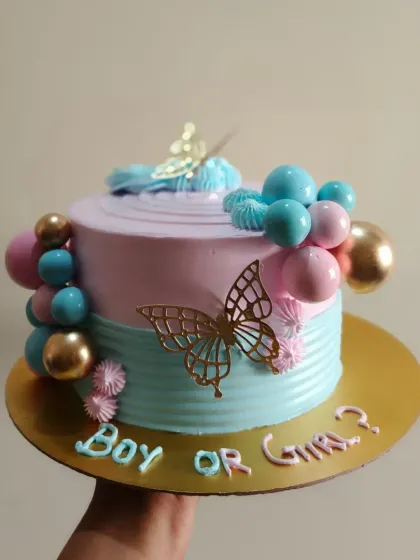 A "Boy or Girl?" gender reveal cake in pastel pink and blue, decorated with golden butterflies and decorative spheres.