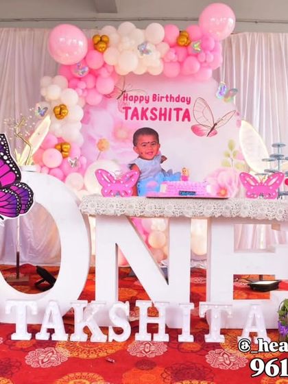 A first birthday setup combining butterfly and fairy elements, featuring a large "ONE" table for the cake. The decor includes fairy wing cutouts, a personalized backdrop, and a pink and white balloon arrangement.