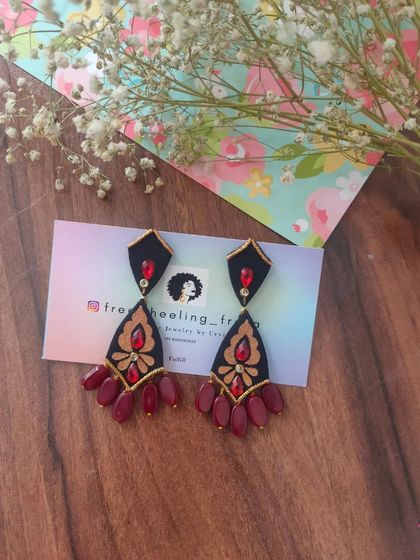 A pair of elegant, hand-painted chandelier earrings. The black fabric base is adorned with golden and red painted details and finished with maroon bead drops.