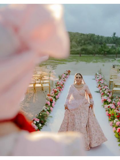 The bride's grand entrance. Walking down an aisle lined with flowers on the sand, her expression is a beautiful mix of excitement and grace. The beach setting makes this classic moment feel fresh and magical.