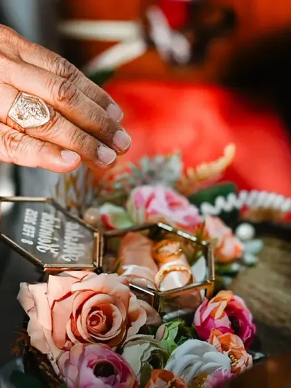 A "multi-layered" detail shot, capturing a hand blessing the wedding rings, which are beautifully displayed in a glass box.