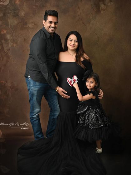 A classic family maternity portrait against a dark, painterly backdrop. The daughter holds tiny shoes to her mother's belly, a touching gesture that makes this photo extra special.