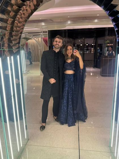 Our international clients looking sharp for an Indian wedding. He is wearing a black Indo-Western sherwani, and she is in a sparkling navy blue crop-top lehenga.