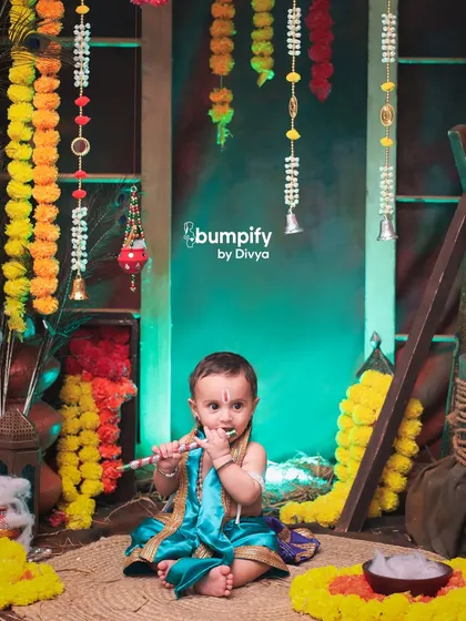 A sweet moment as our little Krishna curiously tastes his flute. The colorful, festive background with marigold garlands adds to the charm of this Janmashtami shoot.