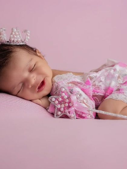 A sleeping baby girl wearing a delicate pearl crown and a pink lace outfit holds a star wand, looking like a tiny princess.