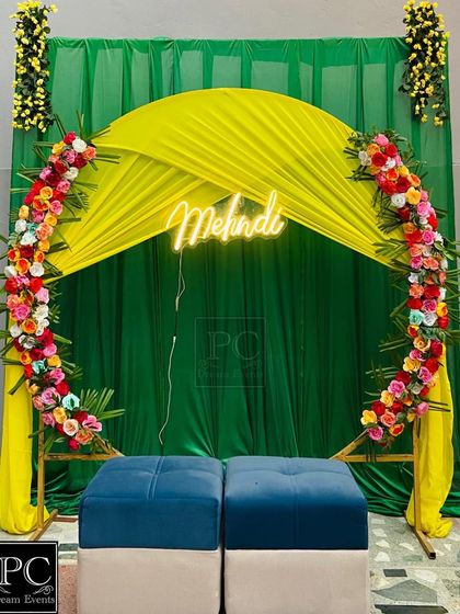 A beautiful Mehndi photo corner with a semi-circular floral arch against a green and yellow draped background. The 'Mehndi' neon sign adds a modern touch.