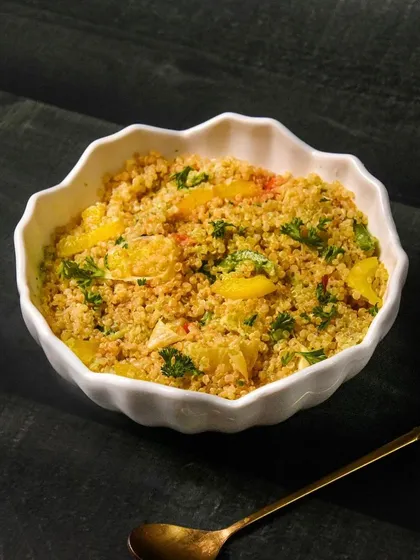 A bowl of healthy quinoa salad with yellow bell peppers and parsley, served with a golden spoon. The dark background makes the light-colored salad stand out.