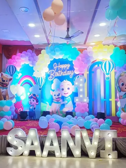 A colorful Cocomelon themed first birthday party. The stage is filled with characters from the show, a rainbow, and pastel balloons, along with large marquee letters for the name "SAANVI."