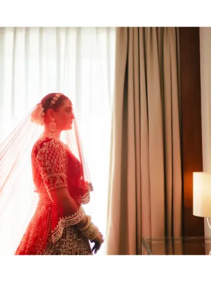 A silhouette of the bride in her red lehenga, standing against a window with her veil catching the light. This artistic shot is a quiet, contemplative moment from her getting-ready story.