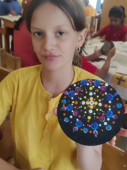 A student from the Inodai Waldorf School displays the beautiful dot mandala she painted on a circular coaster. Her design features a delicate explosion of blue, pink, and white dots.