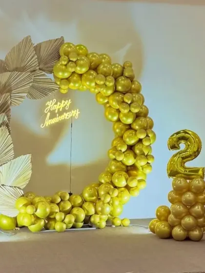 A unique and stylish 25th anniversary backdrop featuring an all-gold balloon arch accented with dried palm leaves. The setup includes a neon sign and a matching balloon column with foil numbers.