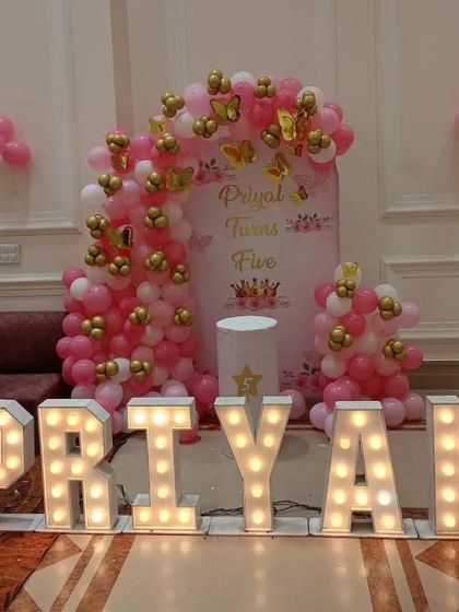 A princess-themed 5th birthday party with large light-up letters spelling the name "PRIYAL". The setup includes a pink and gold balloon arch and a personalized backdrop.