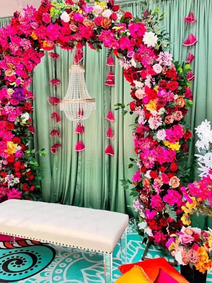 A stunning Mehandi seating with a dense floral arch of pink and red flowers. A small chandelier hangs in the center, adding a touch of elegance to this vibrant setup.