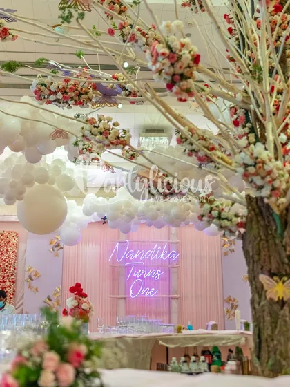 A magical first birthday setup with a 'Nandika Turns One' neon sign, balloon clouds on the ceiling, and a beautiful wishing tree.