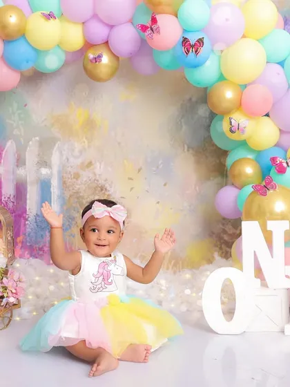A baby girl celebrates her first birthday with a joyful cake smash. Dressed in a colorful rainbow tutu, she smiles brightly amidst a pastel-colored setup with a balloon arch, embodying the fun and excitement of a milestone celebration.