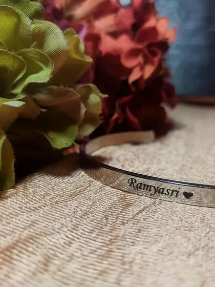 A silver cuff bracelet engraved with a name and a heart symbol, 'Ramyasri ♡', resting near flowers. This shows how you can combine names with symbols for a personal touch.